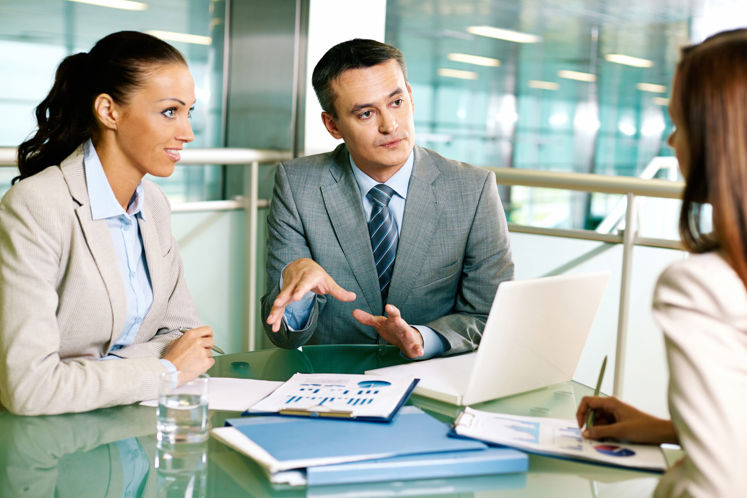 Financial professionals in a collaborative meeting discussing wealth management strategies. A male advisor gestures thoughtfully while explaining concepts. Financial documents with charts and graphs are spread across the glass conference table in a modern office setting, depicting the professional consultation needed when exploring Private Placement Life Insurance options.
